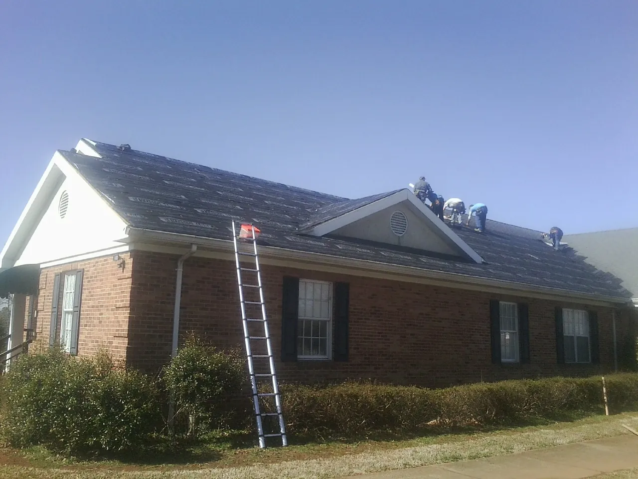 Crew tearing shingles off a brick ranch roof with a ladder set — Emergency Roof Repair work by our crew in Cabin John